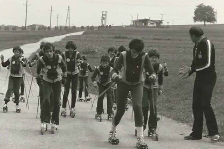 Kinder und Jugendliche beim Skirollertraining auf einer Straße, betreut von einem Trainer.