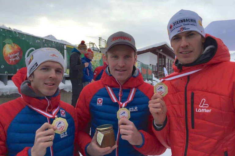 Drei Langläufer mit Medaillen stehen vor einer Schnee-losen Kulisse in Saalfelden.